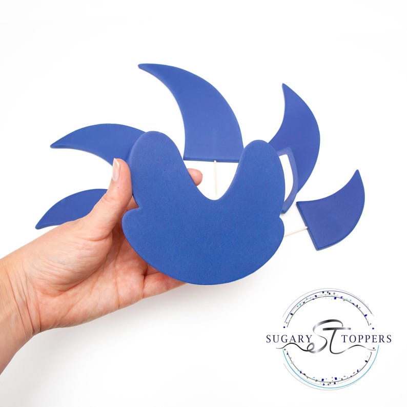 May include: A blue decorative cake topper with a unique design. The topper features several curved shapes and is held by a hand against a white background. The logo for Sugary Toppers is in the bottom right corner.