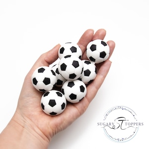 Pu&ograve; includere: Una collezione di piccole decorazioni sferiche a forma di pallone da calcio con il classico motivo a pentagoni bianchi e neri. Questi oggetti rotondi presentano una finitura liscia e sono raggruppati, mettendo in risalto le dimensioni uniformi e il design grafico dettagliato.