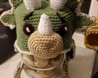 May include: Green crocheted dinosaur hat with white horns and a brown nose. The hat has a long green and white crocheted string attached.