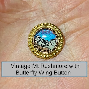 May include: A circular button featuring a gold-toned scalloped metal border surrounding a central inlay. The inlay displays a graphic of the Mount Rushmore monument set against a vibrant, iridescent blue butterfly wing background.