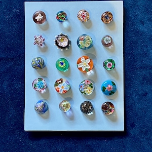 May include: A collection of twenty-one colorful glass cabochons, each with unique designs. The cabochons are arranged on a light blue surface, showcasing a variety of floral, abstract, and patterned elements. The cabochons are round and vary in size.