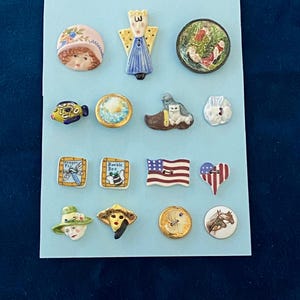 Op de afbeelding: Een verzameling decoratieve keramische knopen met diverse ontwerpen, waaronder een engel, een hommel en een Amerikaanse vlag. De knopen zijn in verschillende vormen en maten, met een mix van kleuren en details.