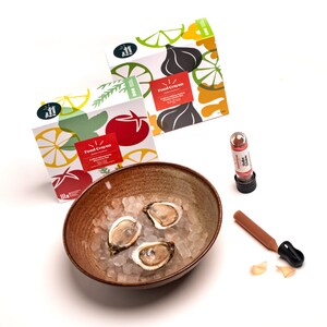 May include: A bowl of ice with three oysters, a small brown bottle with a red cap, and a brown and orange food crayon with the text "Food Crayon" on the packaging. The packaging is colourful and features illustrations of lemons, tomatoes, and herbs.