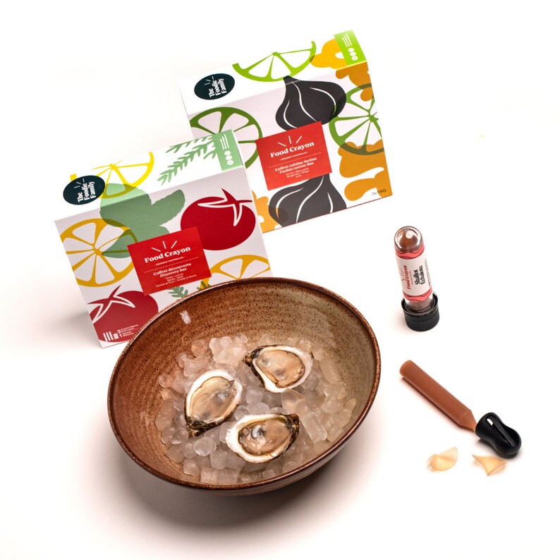 May include: A bowl of ice with three oysters, a small brown bottle with a red cap, and a brown and orange food crayon with the text "Food Crayon" on the packaging. The packaging is colourful and features illustrations of lemons, tomatoes, and herbs.