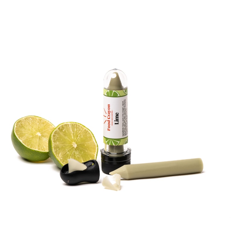 Puede incluir: Un cray&oacute;n alimenticio de color verde lima en un tubo transparente con una tapa negra. El tubo tiene una etiqueta que dice "Food Crayon" y "Lime". Hay dos mitades de lima al lado del tubo y un trozo de cray&oacute;n roto.