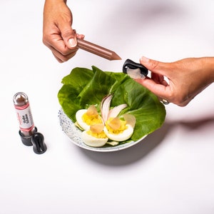 May include: A white plate with a green lettuce base, three deviled eggs, a red onion slice, and a small black and brown bottle with the text "Sushi Essence" on the label. A hand is holding a brown stick with a black cap.