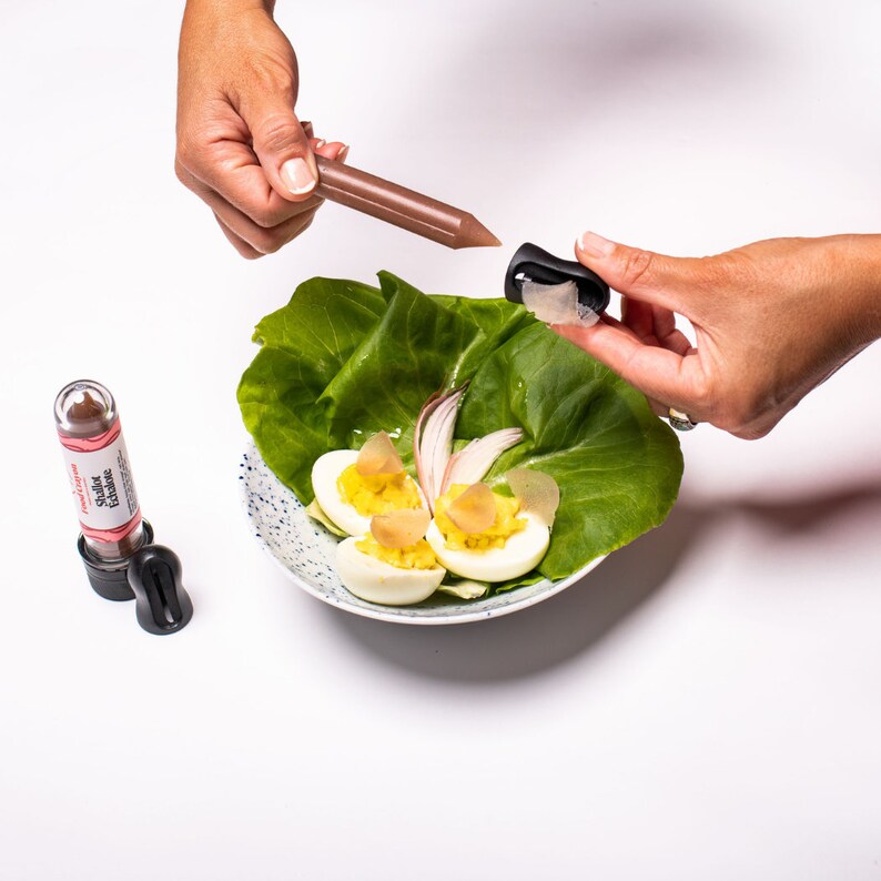 May include: A white plate with a green lettuce base, three deviled eggs, a red onion slice, and a small black and brown bottle with the text "Sushi Essence" on the label. A hand is holding a brown stick with a black cap.