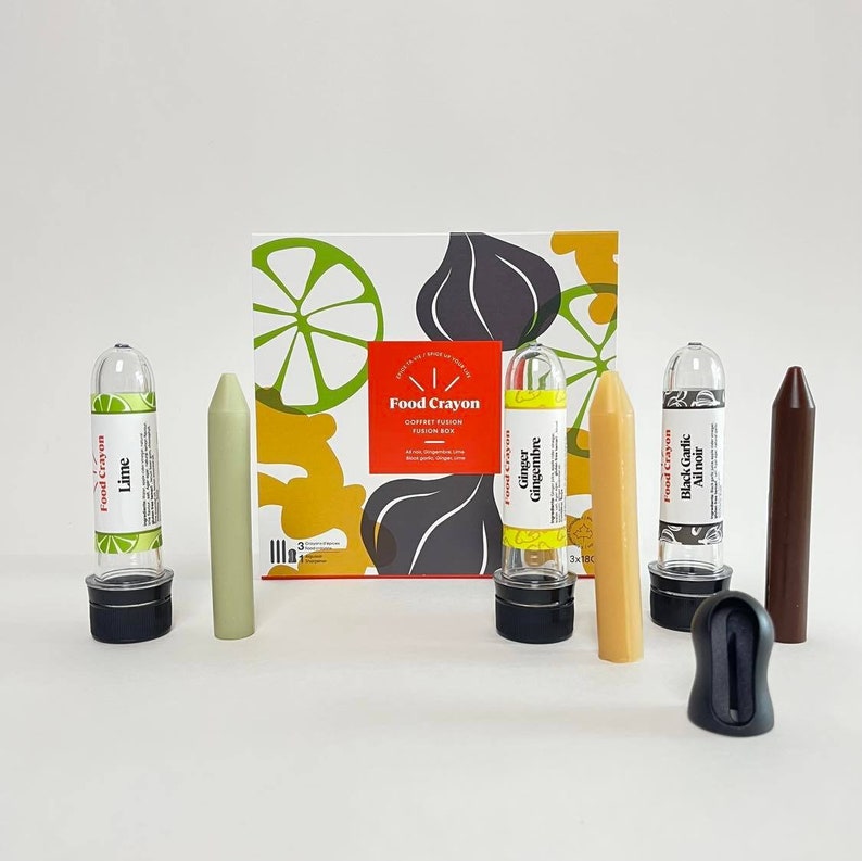 Puede incluir: Un juego de seis crayones comestibles en tubos transparentes con etiquetas para lima, jengibre y ajo negro. Los crayones est&aacute;n dispuestos sobre una superficie blanca con una caja en el fondo. La caja tiene un dise&ntilde;o de limas, jengibre y ajo.