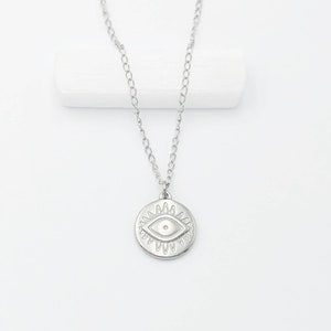 May include: Silver necklace with a round pendant featuring a stylised eye design. The eye is surrounded by a circle of points.