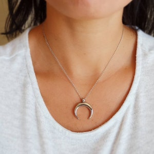 Puede incluir: Un collar con un colgante de media luna de plata en una cadena de plata. El collar lo lleva una persona con el pelo oscuro.
