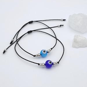 Può includere: Due braccialetti in corda nera con perline argentate e ciondoli blu a forma di occhio turco. Un ciondolo è blu chiaro e l'altro è blu scuro.