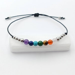 Può includere: Braccialetto regolabile con sette perline colorate di pietre preziose che rappresentano i chakra. Le perline sono disposte su un cordoncino nero, con perline di metallo color argento su ogni lato. Il braccialetto è esposto su una superficie bianca.