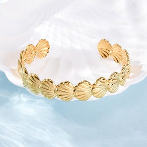 Peut inclure: Un bracelet manchette doré avec un motif de coquillage. Le bracelet est composé d'une série de coquillages détaillés en forme d'éventail reliés entre eux. La conception ouverte permet de le porter facilement. Le bracelet est présenté sur une surface blanche avec un fond bleu clair.