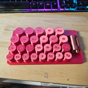 Puede incluir: Una herramienta roja impresa en 3D con múltiples piezas cilíndricas de varios tamaños, cada una etiquetada con un número. La herramienta está diseñada para medir o sujetar objetos. Los números van del 4 al 16,5.