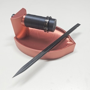 May include: A copper-colored tool with a black cylindrical component and a black, flat, pointed tool. The copper-colored base has a curved design. The cylindrical component has a ring around it. The tools are on a white surface.