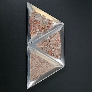 May include: A top-down view of a geometric arrangement of three triangular metal trays. Each tray contains a different type of light brown, textured material, possibly flakes or granules, against a black background.