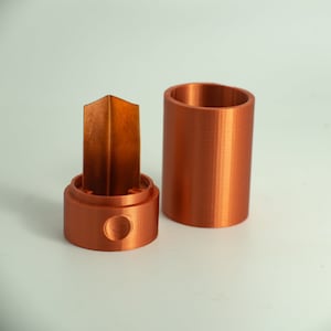 May include: A copper-colored 3D printed container with a removable lid and a small, copper-colored, 3D printed insert. The container is cylindrical and the insert is shaped like a small, thin, rectangular prism.