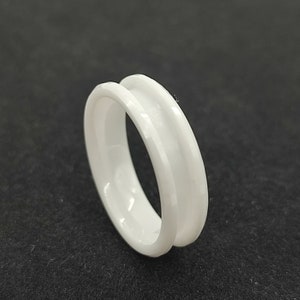May include: A white ceramic ring with a wide, flat band and a beveled edge. The ring is simple and elegant.