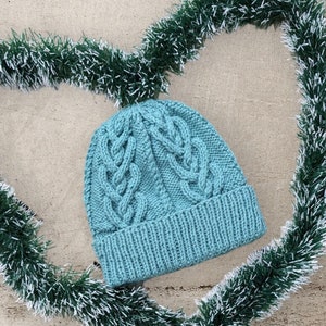 May include: A teal-colored knitted beanie hat with a cable knit design. The hat has a ribbed brim and a textured pattern on the crown. The hat is surrounded by a green and white garland in a heart shape.
