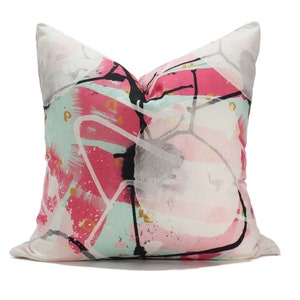 May include: A white cushion with a pink, blue and black abstract design. The design features bold lines and splashes of colour.
