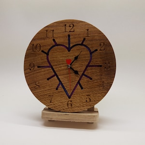 May include: A wooden clock with a heart-shaped cutout in the center. The clock face has Roman numerals and a red second hand. The clock is on a small wooden stand.