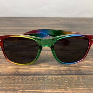 May include: Rainbow-coloured sunglasses with dark lenses. The frame is made of plastic and has a gradient of red, orange, yellow, green, blue, and purple.