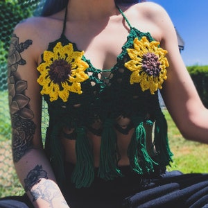 May include: A green crochet halter top with sunflower embellishments and fringe. The top is made of a soft, lightweight yarn and has a relaxed fit.