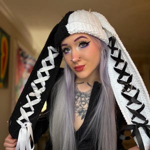 May include: A split-color crochet hat featuring long, dangling ear flaps. The left side is black with white ribbon lacing, while the right side is white with black ribbon lacing. Both sides feature silver-toned heart-shaped metal rings.