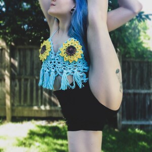 May include: A woman with blue hair is wearing a blue crocheted top with sunflowers and black shorts. She is doing a leg split.