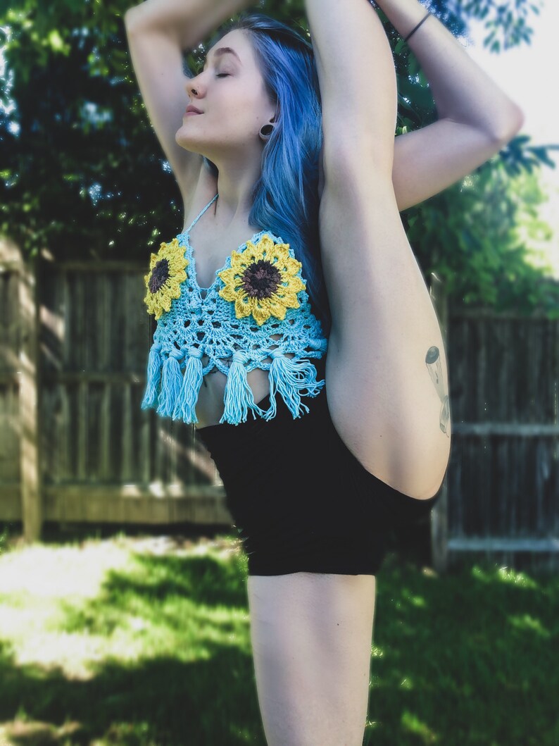 May include: A woman with blue hair is wearing a blue crocheted top with sunflowers and black shorts. She is doing a leg split.