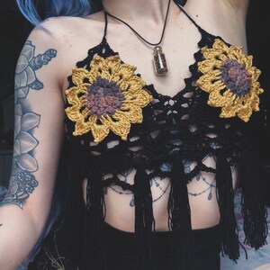 May include: A black crochet halter top with sunflower-shaped crocheted embellishments and fringe details. The top features a small glass vial pendant on a black cord.