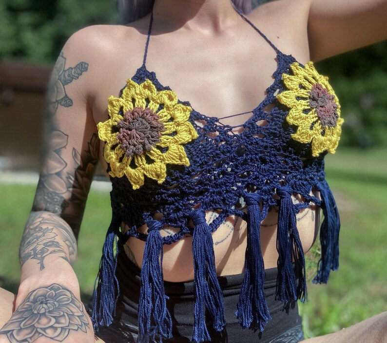 May include: A navy blue crochet top with sunflower-shaped embellishments. The top has fringe detailing and a halter neck style.