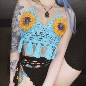 May include: A blue crocheted halter top with sunflower embellishments and tassels. The top is worn over a black crocheted bikini bottom.
