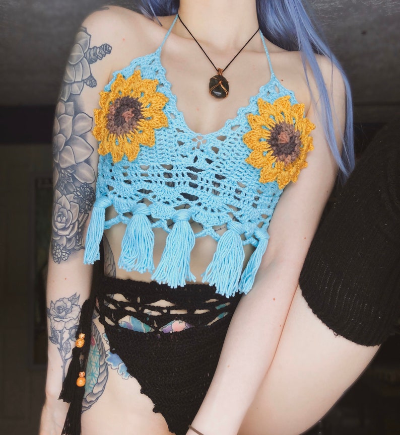 May include: A blue crocheted halter top with sunflower embellishments and tassels. The top is worn over a black crocheted bikini bottom.