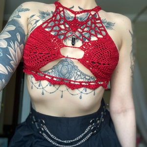 May include: Red crochet halter top with a spiderweb design and a black gemstone pendant. The top has a cutout in the front and is worn over a black skirt with a silver chain belt.