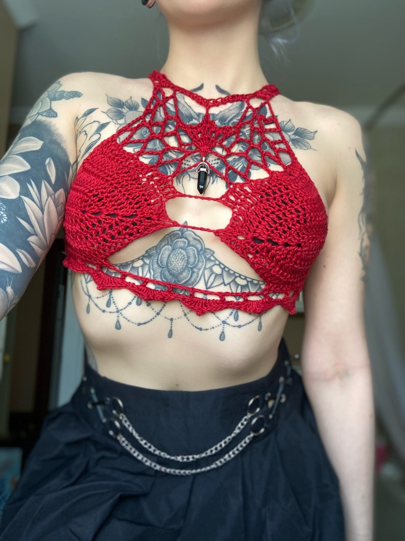 May include: Red crochet halter top with a spiderweb design and a black gemstone pendant. The top has a cutout in the front and is worn over a black skirt with a silver chain belt.