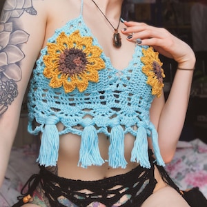 May include: A blue crochet halter top with sunflower details and tassels. The top is made with a delicate crochet stitch and features a light blue color with yellow and brown accents.