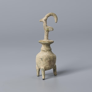 Puede incluir: Un recipiente de bronce antiguo y desgastado con un diseño único. El recipiente tiene una base bulbosa sostenida por tres patas cortas. Un tallo delgado se eleva hacia una parte superior ensanchada, y un elemento curvo en forma de cuerno se extiende hacia arriba. El color general es un bronce apagado y envejecido.