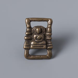 May include: A bronze-colored metal ornament featuring a seated Buddha figure within a rectangular frame. The Buddha is in a meditative pose, and the frame has a handle at the top and a slot at the bottom. The piece has an aged, antique appearance.