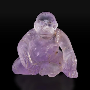 Puede incluir: Una pequeña figurita de amatista tallada que representa una figura sentada con una cara redonda y ojos grandes. La figurita es de color púrpura claro y tiene un acabado liso y pulido.