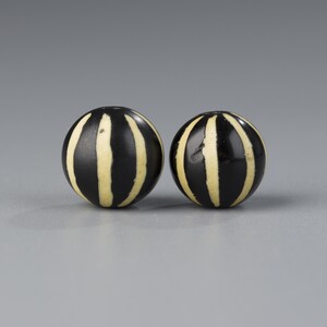 May include: Two spherical beads featuring a smooth black surface with vertical cream-colored stripes. Each bead has a rounded shape and a consistent pattern of alternating dark and light bands that wrap around the circumference of the object.