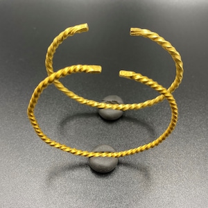 May include: Two gold-toned twisted bangle bracelets. The bracelets are stacked on top of each other and are resting on a gray surface.