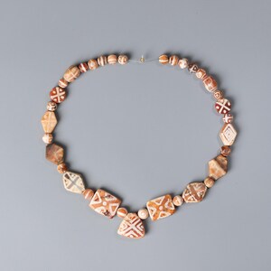 Puede incluir: Un collar con cuentas marrones y beige de varias formas, incluyendo redondas, de diamante y cuadradas, con patrones geométricos blancos. El collar está ensartado en un cordón fino y transparente y se muestra sobre un fondo gris.