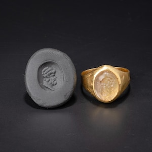 May include: A gold ring with an oval-shaped, light-colored stone featuring a carved bust. Beside it, a dark gray oval seal with a matching bust carving. Both items are set against a dark background.