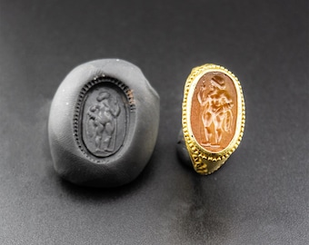 May include: A gold ring with an oval carnelian stone featuring a carved image of a nude figure holding a staff. The ring is set in a gold bezel with a beaded edge.