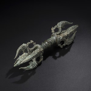 May include: A bronze-coloured vajra, a ritual object with three prongs on each end, and a central handle. The vajra has intricate details and a weathered appearance, suggesting age. The background is a solid black.