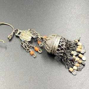 Op de afbeelding: Een zilveren oorbel met een hangende bedel met een kleine, ronde, zilveren kraal met een getextureerd oppervlak. De bedel is bevestigd aan een ketting met meerdere kleine zilveren kralen en een paar oranje kralen. De oorbel is ook versierd met een zilveren haak.