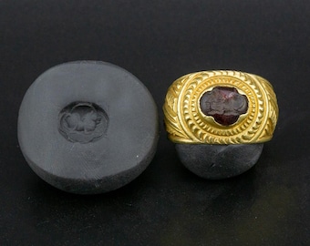 May include: A gold ring with a red gemstone set in a bezel. The ring is decorated with a detailed design around the gemstone. The ring is shown next to a mold of the ring.