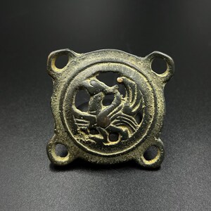 May include: A bronze pendant with a circular design featuring a stylized bird or animal. The pendant has four loops on the edges, suggesting it was once part of a larger piece of jewelry.