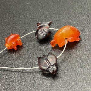 May include: Four carved amber beads, two orange and two dark brown, strung on a thin silver wire. The beads are shaped like animals, including a fish, a pig, and two beetles.